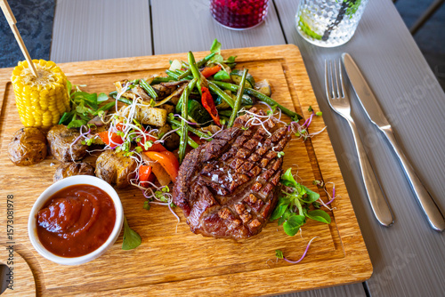 Grilled pork steak, vegetable, maize, green leaf, sauce, cutlery , drink on wooden board.