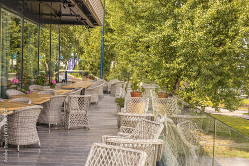 Empty outdoor café terrace with wicker furniture, wooden tables, flowers, and vibrant green trees creating a cozy dining environment in a natural setting.
