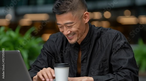 Asian man with short hair, wearing a black shirt, is smiling while working on a laptop in a modern workspace, surrounded by greenery and soft lighting, showcasing a positive atmosphere