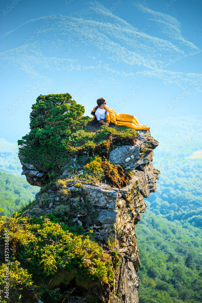 Obraz premium Man relaxing in sleeping bag on mountain cliff, enjoying scenic sunrise view over forested hills