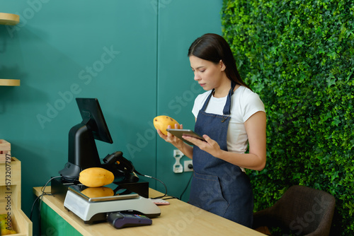 Caucasian female business owner using tablet device while checking mangoes before placing on scale during fruit inventory preparation behind grocery counter in modern small shop setup before opening