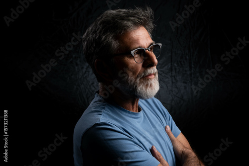 Photos Handsome mature man sitting with crossed arms, side profile, mysterious gaze on black background, chiaroscuro lighting mood