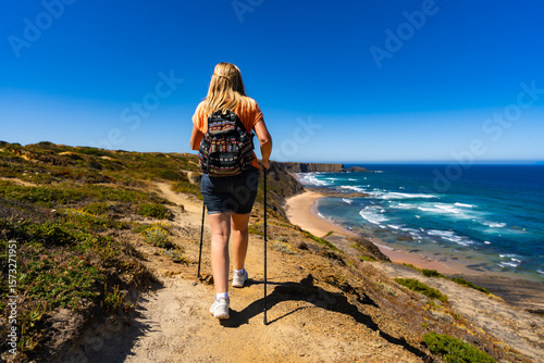 Beautiful mid adult woman hiking on tourist trail along atlantic ocean in nature park Vicentine Coast in Portugal on sunny summer day. Back view	