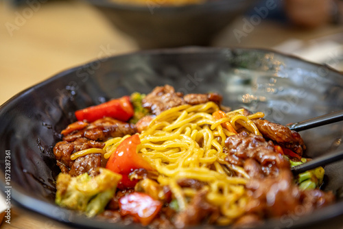 Close-up of stir-fried noodles with beef, bell peppers, and vegetables in a black bowl, served with chopsticks. Asian fusion cuisine, vibrant and appetizing.
