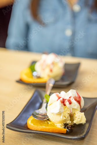 Close-up of vanilla ice cream served with whipped cream, berry sauce, orange slice, and cherry on a black plate at an Asian restaurant. Elegant and refreshing dessert presentation.