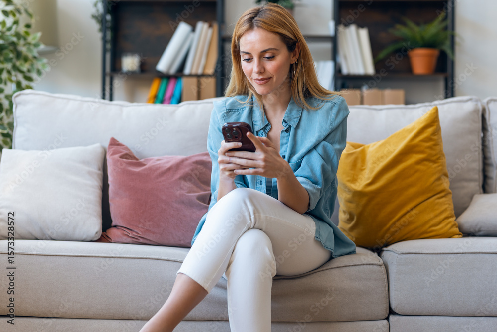 Obraz premium Beautiful young woman using her mobile phone sitting on couch at home