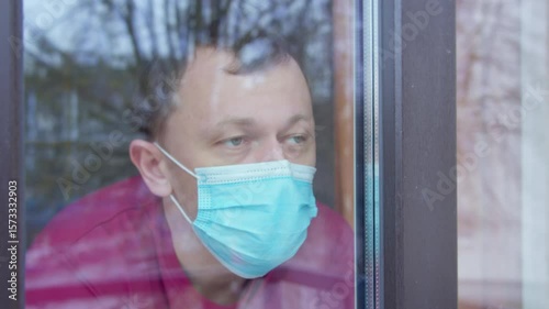 Man in medical mask, sitting at home looking at the street through the window, reflection of the street