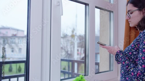 Woman stands by the window waiting, reading a message on her phone