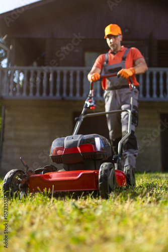 Gardener mowing the lawn in front of a wooden house during a sunny day