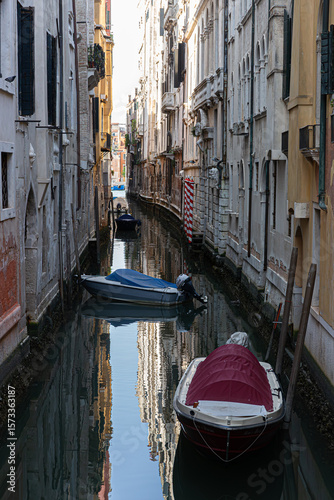  Boats with colorful covers are moored in a quiet, narrow canal, with their reflections perfectly mirrored in the dark, still water.