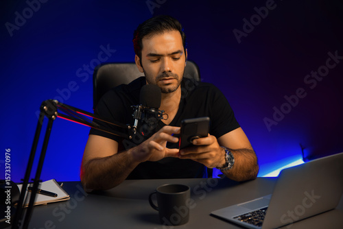 Broadcaster talking to listener on live streaming online, checking message from smartphone on social media in host channel by creative positive thinking concept at purple neon light studio. Surmise.