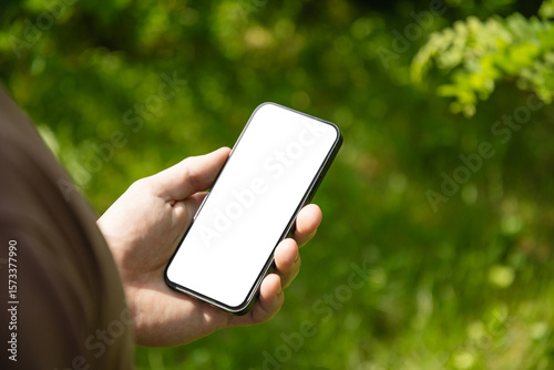 Mans hands holding smartphone with white empty screen in outdoor green setting. Check apps, focused on device, chatting, texting. Fitness progress on app. Side view. Close up.