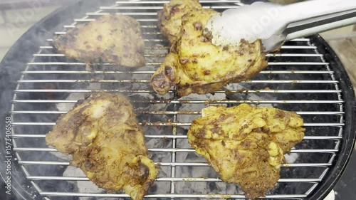 Closeup of tongs turning pieces of chicken, cooking on a grate over hot charcoal in an outdoor barbecue grill.