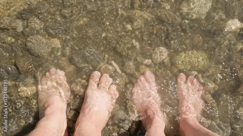 Pair of women's and men's barefoot feet are washed by wave of water from pond standing on pebble beach