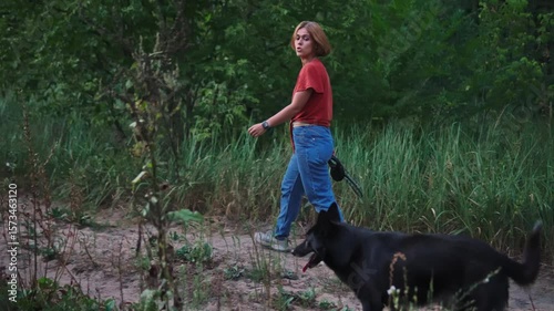 Woman walks on forest footpath in the early summer evening holding leash of her playful black dog. Slowmo of smiling young adult lady going in woodland with her pet. Friendship of animal and person