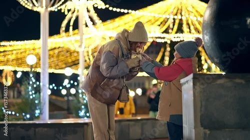 Mother and daughter stand at a festive Christmas fairground at a cold evening and share a shawarma a shawarma sandwich together enjoying a tasty snack in the chilly night air among holiday decorations