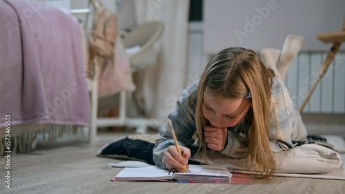 Concentrated schoolgirl in casual clothes is lying on the floor and write in copybook with a pen filmed in slow motion. Caucasian kid is doing homework in cozy home atmosphere making notes in a book