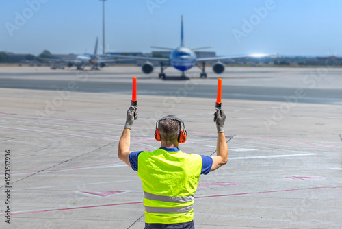 Aircraft marshaller in PPE guiding airplane on runway with marshalling wands at airport.