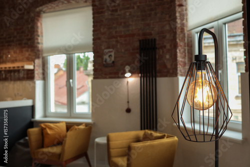 Close-up of an industrial-style floor lamp with a glowing Edison bulb in a metal cage frame, set in a cozy living room with mustard armchairs, exposed brick walls, and large windows