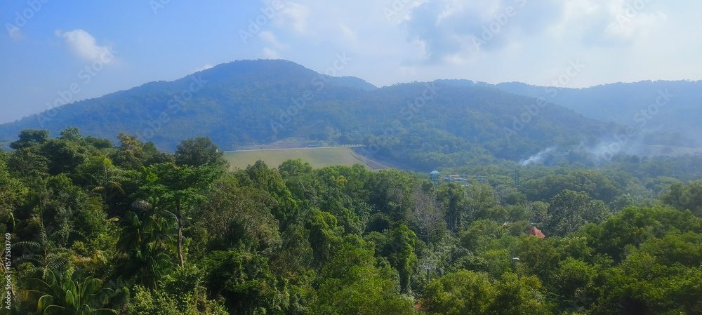 Fototapeta premium Panoramic view of rolling, tree-covered mountains and dense, vibrant green jungle. The clear sky highlights the vastness of the natural landscape, with only subtle signs of human presence.