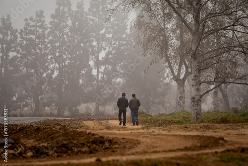 Two men walking on a dirt road on a foggy morning with trees in the background
