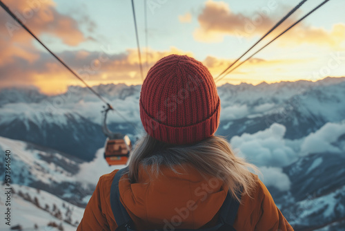 Rear View of Woman in Gondola at Sunset. A woman in a red beanie rides a gondola lift at sunset, overlooking snowy mountains and glowing clouds.