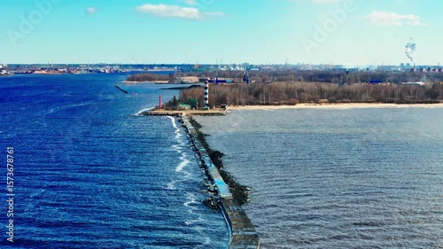 Wallpaper Mural A striped lighthouse stands on a peninsula with a long breakwater stretching between river and sea. Coastal landscape with distant city skyline under blue sky. Torontodigital.ca