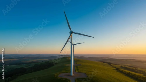 Wind Turbines Stand Tall At Sunset On Green Hillside With Hilly Terrain Under Clear Blue Sky