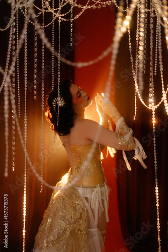Brunette young woman, Caucasian, dressed as vintage cabaret dancer, posing front view under hanging beads on dark background. Concepts: cabaret, retro glamour, burlesque, stage show.