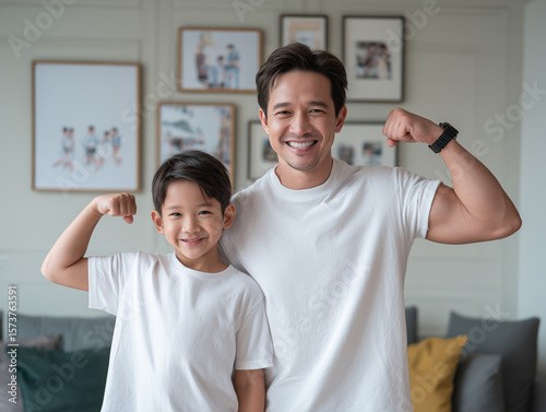 young boy and his father flex their muscles, both smiling joyfully in matching white t shirts. cozy living room features family photos in background, creating warm atmosphere