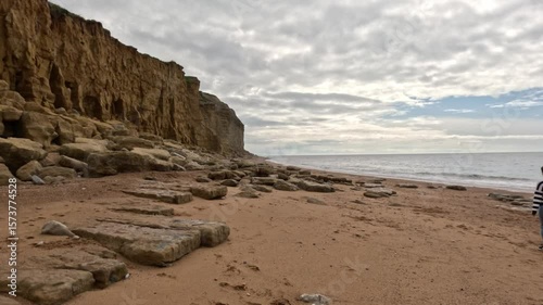 The camera moves sideways to the right as the female walks away along the beach wide the sea on the right and the cliffs left. A women walking along a Jurassic Coast beach in Dorset.