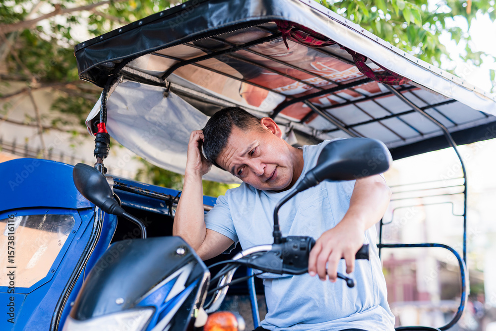Obraz premium Asian man wearing a blue shirt looks irritated and disappointed while scratching his head, clearly frustrated with his tricycle’s motor not starting.