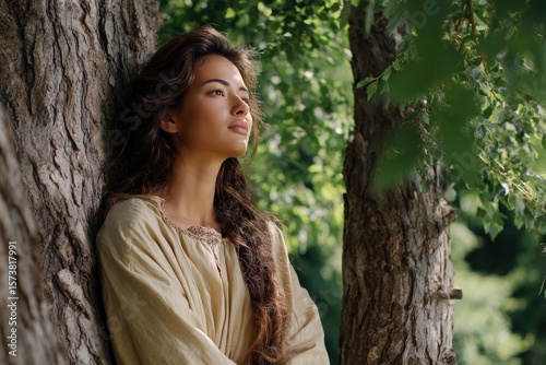 Woman in peasant blouse leans against a roughbarked tree gazing upward