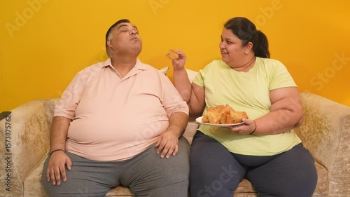 Overweight obese indian couple enjoy fried samosa at home. Plus size husband and wife eating unhealthy junk food together. diabetes, healthcare obesity.4k