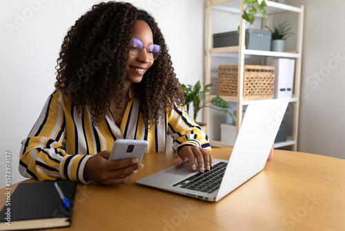 Happy African American woman using phone and laptop working at home office.