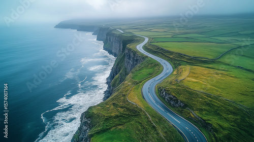 Winding Coastal Road Along Green Cliffs. Aerial view of a scenic winding road along steep green cliffs above the ocean, with waves crashing below and mist in the distance.