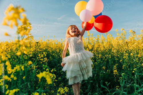 Girl in a white dress holding colorful balloons in a rapeseed field