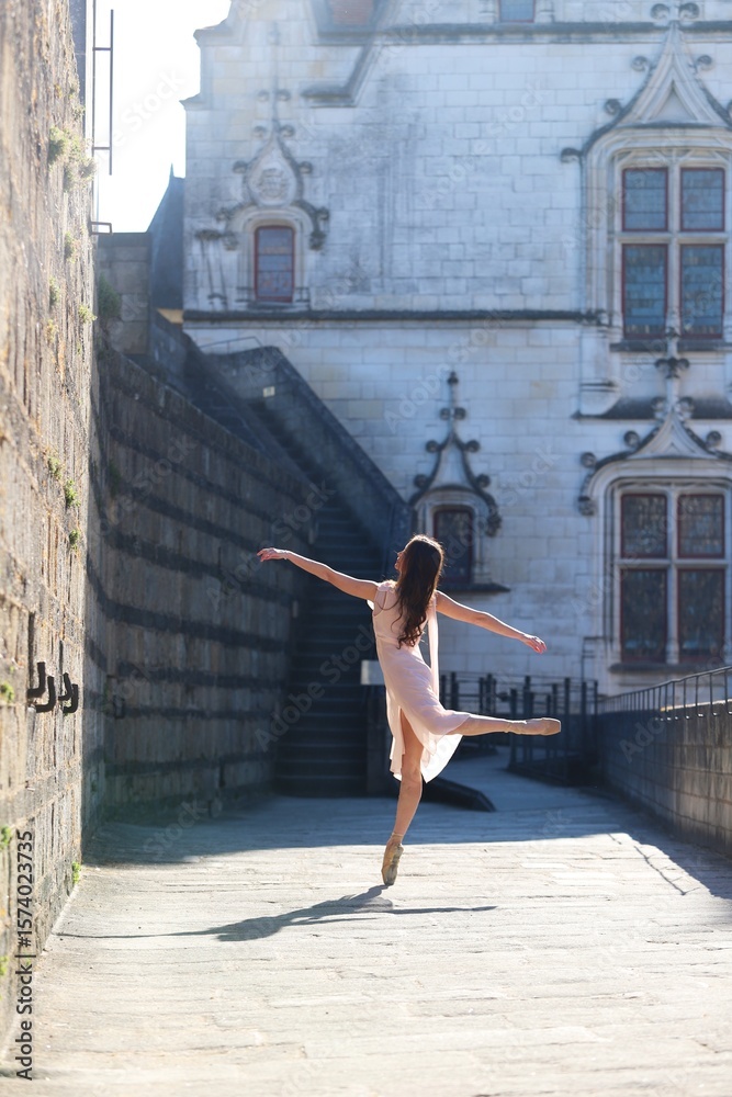 Fototapeta premium Graceful ballerina in soft pink dress dancing on pointe in sunlit alley with historic French castle in the background