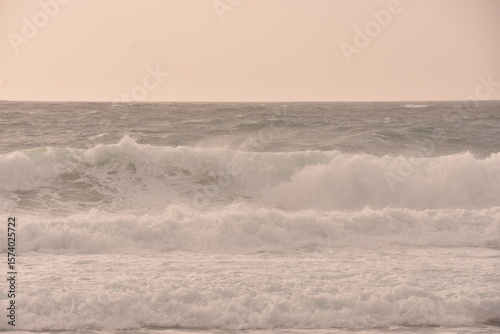 Fototapeta Naklejka Na Ścianę i Meble -  Powerful ocean waves crashing under a soft pastel-toned sky during sunset. The golden light of the setting sun casts a warm glow over the scene, creating an atmosphere that is both serene and dramatic