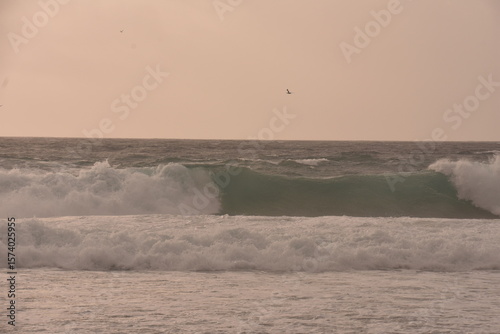 Fototapeta Naklejka Na Ścianę i Meble -  Powerful ocean waves crashing under a soft pastel-toned sky during sunset. The golden light of the setting sun casts a warm glow over the scene, creating an atmosphere that is both serene and dramatic