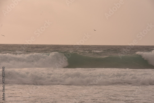 Fototapeta Naklejka Na Ścianę i Meble -  Powerful ocean waves crashing under a soft pastel-toned sky during sunset. The golden light of the setting sun casts a warm glow over the scene, creating an atmosphere that is both serene and dramatic