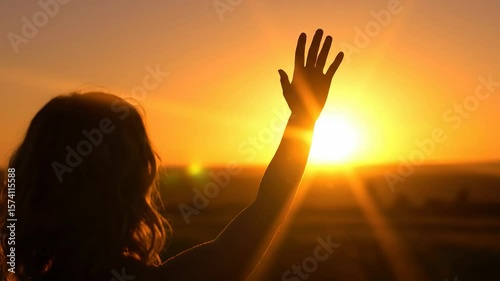 A backlit figure raises their hand towards a brilliant setting sun