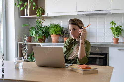 Pensive exhausted woman work on laptop studying online with books and water glass at home. Freelancing, digital nomad, remote learning, video calls, self education, flexible work, modern professional
