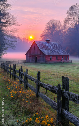 Beautiful sunrise over a rustic barn. A serene sunrise casts golden light over a charming red barn surrounded by a misty landscape and wooden fence.