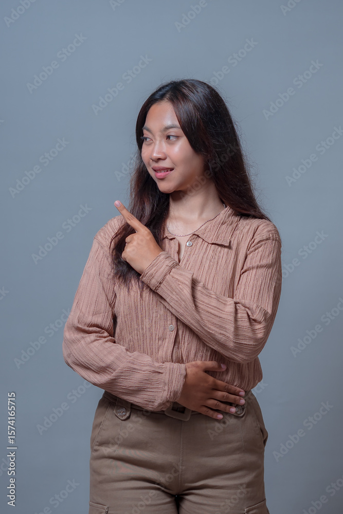 An Indonesian woman in a beige blouse smiling and pointing upward with one hand while the other rests on her waist.expressing a confident and friendly demeanor. An Indonesian woman in a beige blouse smiling and pointing upward with one hand while the other rests on her waist.expressing a confident and friendly demeanor.