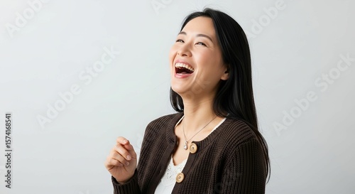 Laughing East Asian Woman in Ribbed Brown Cardigan and Lace-Trimmed White Top with Pendant Necklace on Light Background photo
