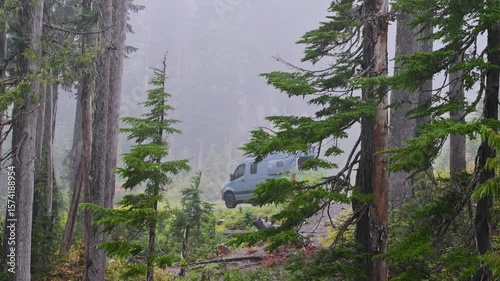 Drone view of a misty forest clearing with a camper van parked along a dirt path among tall pine trees in the wilderness