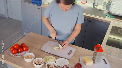 Overhead view of a home cook slicing white onion into thin rings on a cutting board in a bright kitchen. Fresh ingredients, tomatoes, cheese, and seafood are laid out on the counter. 