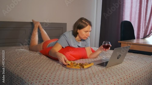 Young woman lying on bed, savoring a slice of pizza with closed eyes, holding a glass of red wine nearby. Laptop open in front of her. Cozy evening alone, enjoying the taste of food. 