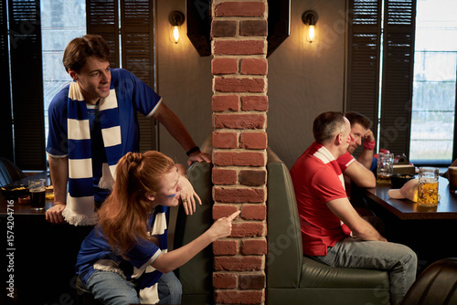 Caucasian young man and woman, wearing blue soccer scarves, leaning around brick wall, playfully interacting with two middle aged men in red jerseys, sitting at table watching football game